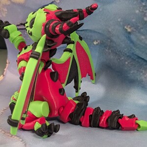 May include: A 3D-printed dragon figurine in a seated pose. The dragon features a vibrant color scheme of red, green, and black. The wings are green with red undersides. The dragon's body is red with green accents and black claws.