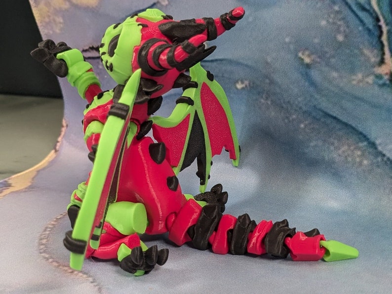 May include: A 3D-printed dragon figurine in a seated pose. The dragon features a vibrant color scheme of red, green, and black. The wings are green with red undersides. The dragon's body is red with green accents and black claws.