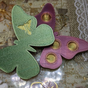 May include: Two decorative butterfly-shaped ornaments with a textured surface. One butterfly is green with a gold butterfly design and the other is pink with three gold circles.