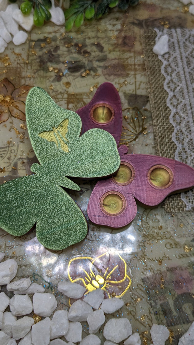 May include: Two decorative butterfly-shaped ornaments with a textured surface. One butterfly is green with a gold butterfly design and the other is pink with three gold circles.
