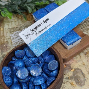 May include: A wooden bowl filled with small, round, blue glitter-covered wax melts. A small wooden block holds two similar wax melts. A card with the text "Sapphire Eclipse" is also present.