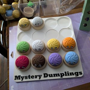 May include: A white tray with twelve colorful, smiling dumpling toys. The dumplings are in various colors, including yellow, green, orange, red, brown, purple, and blue. The tray has the words "Mystery Dumplings" in black.