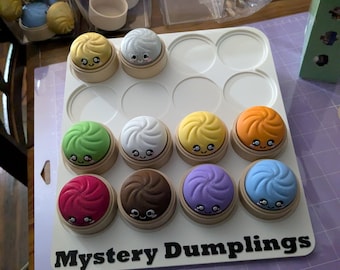 Pode incluir: Uma bandeja branca com doze brinquedos de dumpling coloridos e sorridentes. Os dumplings são de várias cores, incluindo amarelo, verde, laranja, vermelho, marrom, roxo e azul. A bandeja tem as palavras "Mystery Dumplings" em preto.