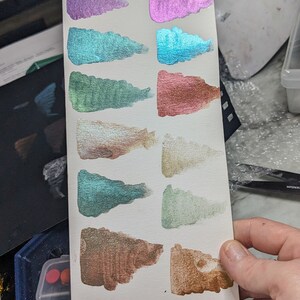 Pu&ograve; includere: Un cartoncino bianco con 12 campioni di vernice iridescente in vari colori, tra cui argento, viola, verde acqua, verde, oro e marrone. Il cartoncino &egrave; tenuto in una mano.