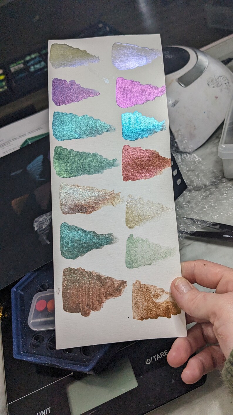 Pu&ograve; includere: Un cartoncino bianco con 12 campioni di vernice iridescente in vari colori, tra cui argento, viola, verde acqua, verde, oro e marrone. Il cartoncino &egrave; tenuto in una mano.
