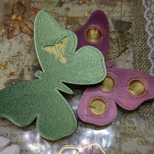 May include: Two resin butterfly ornaments with a textured surface. The butterflies are green and pink with gold accents. The butterflies are on a white background with gold glitter and white stones.