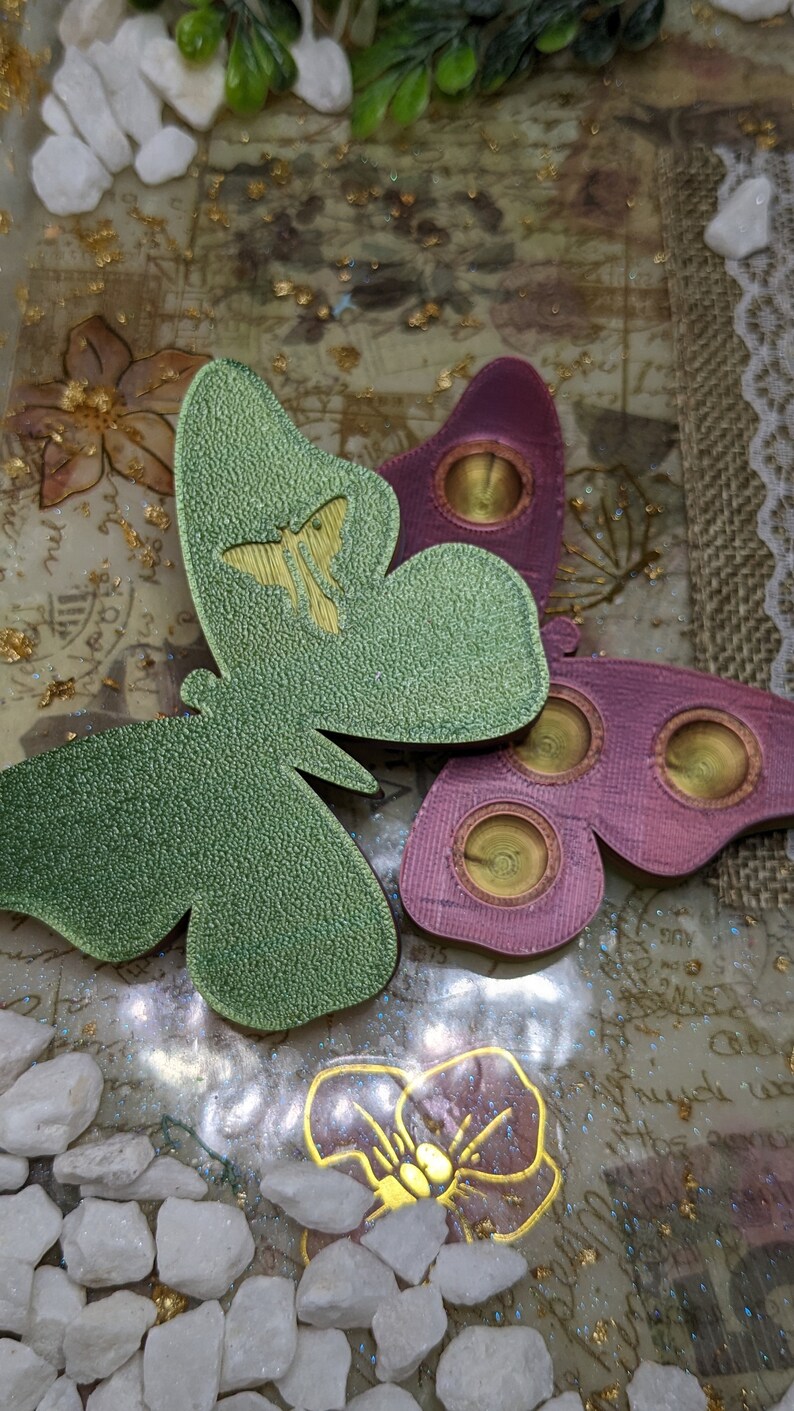 May include: Two resin butterfly ornaments with a textured surface. The butterflies are green and pink with gold accents. The butterflies are on a white background with gold glitter and white stones.