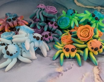 May include: A collection of colorful, hand-painted spider figurines with floral accents. The spiders are made of resin and have a variety of colors, including blue, green, orange, and purple. Each spider has a unique design and is adorned with a flower.
