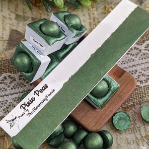 Peut inclure: Une peinture métallisée verte appelée "Pixie Peas" de The Blooming Faerie. La peinture est dans un petit récipient carré avec un puits rond de peinture. Il y a plusieurs petits échantillons ronds de la peinture sur une surface.