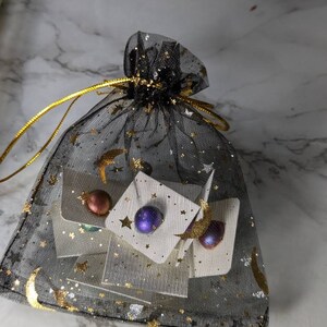 Pu&ograve; includere: Tre paia di orecchini in una borsa di rete nera con stelle e lune dorate. Gli orecchini sono rotondi e hanno una finitura metallica. Un paio &egrave; in rame, un altro &egrave; viola e un altro &egrave; dorato.