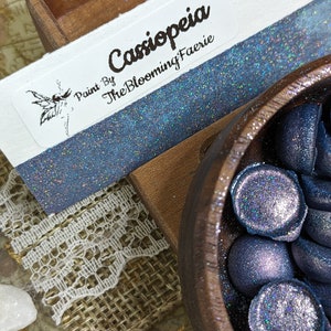 May include: A small wooden bowl filled with blue and purple iridescent glitter resin. The bowl is sitting on a piece of burlap fabric. A label with the text "Paint By The Blooming Faerie" and the name "Cassiopeia" is visible in the background.