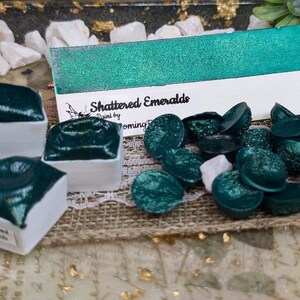 May include: A set of green watercolor paint pans with the label "Shattered Emeralds" and a sample of the paint on a white card. The paint is a shimmery, metallic green.