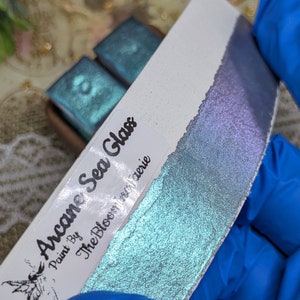 May include: A small sample of Arcane Sea Glass iridescent paint by The Bloom in aerie. The paint is a light blue with a shimmery finish.