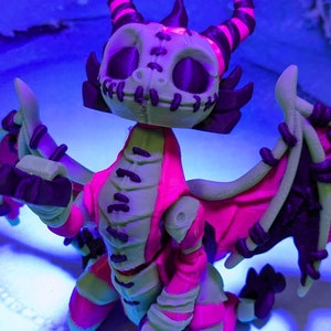 May include: A 3D-printed dragon figurine with a skeletal face, horns, and wings. The dragon features a color scheme of green, pink, and black. The wings are spread, and the figure is set against a blue background.