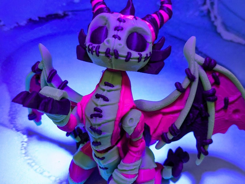 May include: A 3D-printed dragon figurine with a skeletal face, horns, and wings. The dragon features a color scheme of green, pink, and black. The wings are spread, and the figure is set against a blue background.