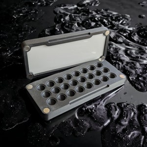 May include: A black plastic case with a hinged lid and 25 round compartments. The case is open and the compartments are visible.