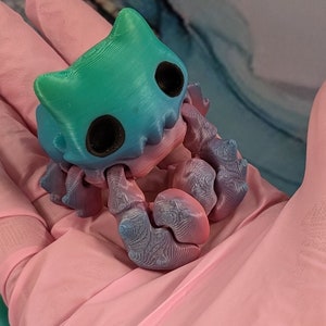 Pode incluir: Uma pequena figura de um caranguejo impressa em 3D, de cor azul esverdeada e azul, com um rosto de desenho animado. O caranguejo est&aacute; sentado em uma superf&iacute;cie rosa.