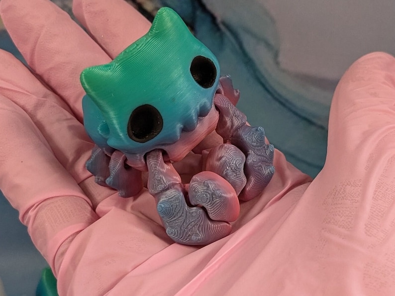 Pode incluir: Uma pequena figura de um caranguejo impressa em 3D, de cor azul esverdeada e azul, com um rosto de desenho animado. O caranguejo est&aacute; sentado em uma superf&iacute;cie rosa.