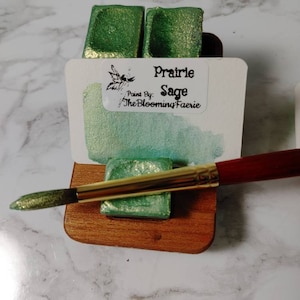 May include: Two small pans of green watercolor paint with a gold shimmer. The pans are labeled "Prairie Sage" and "Paint By: The Blooming Faerie". A wooden paint tray holds the pans and a gold and red paintbrush is resting on the tray.
