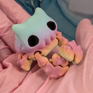 Pode incluir: Um pequeno caranguejo de cor arco-&iacute;ris impresso em 3D com olhos pretos. O caranguejo est&aacute; em pose com suas garras levantadas.