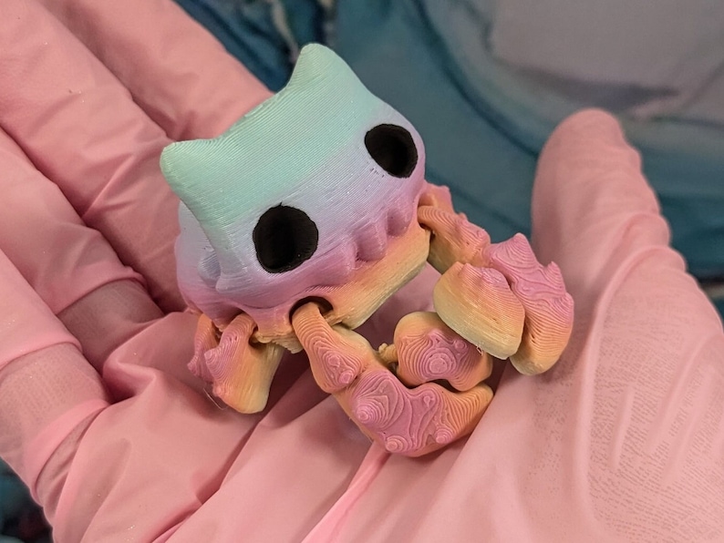 Pode incluir: Um pequeno caranguejo de cor arco-&iacute;ris impresso em 3D com olhos pretos. O caranguejo est&aacute; em pose com suas garras levantadas.