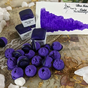 May include: A purple watercolor paint pan with the text "Ultra Marine Violet" and the words "Paint by The Blooming Fox" on a white label. The paint pan is surrounded by small purple beads and white stones on a tray with gold accents.
