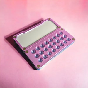 May include: A pink and blue plastic case with a hinged lid and a grid of 25 small round depressions. The case is designed to hold small items, such as pills or jewellery.