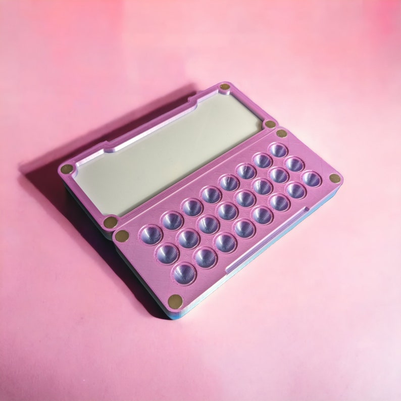 May include: A pink and blue plastic case with a hinged lid and a grid of 25 small round depressions. The case is designed to hold small items, such as pills or jewellery.