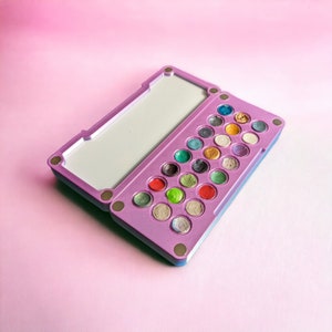 May include: A purple and blue makeup palette with a mirror lid and 24 round, colourful eyeshadows.