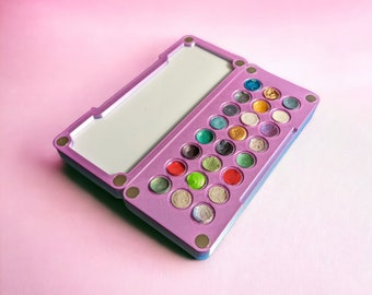 May include: A purple and blue makeup palette with a mirror lid and 24 round, colorful eyeshadows.