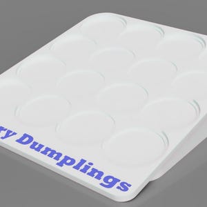 Op de afbeelding: Een witte plastic bak met meerdere cirkelvormige inkepingen, ontworpen om kleine items in te bewaren. De woorden "Mystery Dumplings" zijn in het blauw op de rand van de bak gedrukt. De bak is schuin, met een verhoogde zijkant.