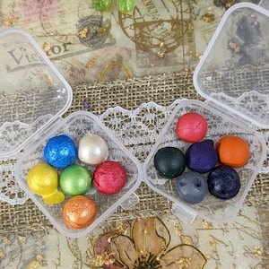 May include: Colorful, round wax melts in clear plastic containers. The melts are various colors, including blue, green, red, and orange. The background includes a floral design and gold leaf accents.