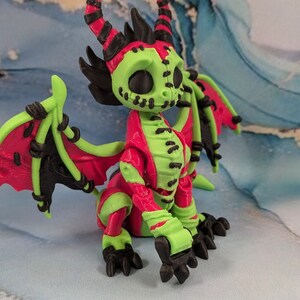 May include: A vibrant, stylized dragon figurine with a unique color scheme. The dragon features a lime green head, limbs, and wings, contrasted by a red body and wings. Black accents and stitching details add to its distinctive appearance.