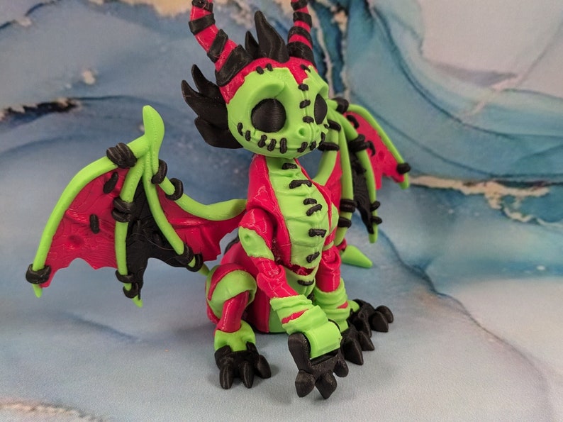 May include: A vibrant, stylized dragon figurine with a unique color scheme. The dragon features a lime green head, limbs, and wings, contrasted by a red body and wings. Black accents and stitching details add to its distinctive appearance.