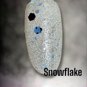 May include: A white nail polish with a clear base and blue and silver glitter. The polish is called "Snowflake" and is part of the "Bloom Dip" collection.