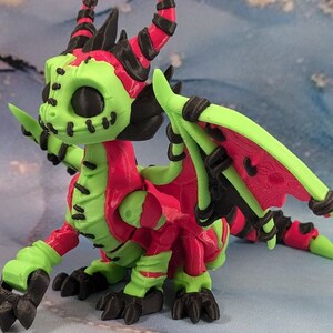 May include: A 3D-printed dragon figurine in vibrant colors. The dragon features a lime green body with red accents on the wings, back, and head. Black details include the horns, claws, and wing supports. The dragon has a stitched-mouth design.