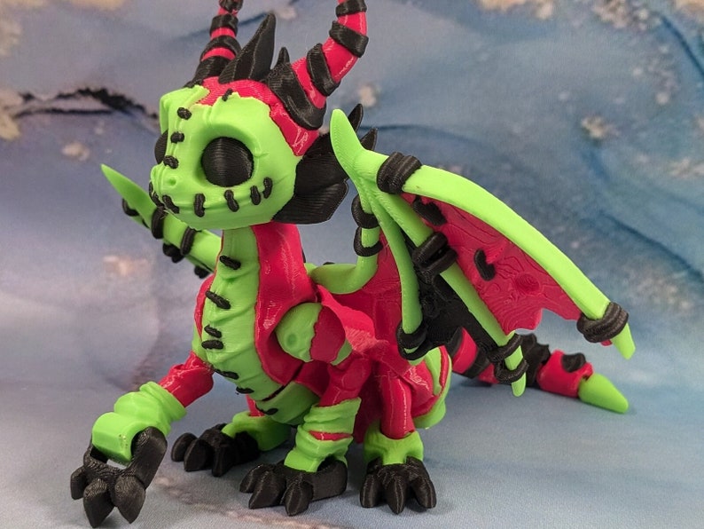 May include: A 3D-printed dragon figurine in vibrant colors. The dragon features a lime green body with red accents on the wings, back, and head. Black details include the horns, claws, and wing supports. The dragon has a stitched-mouth design.