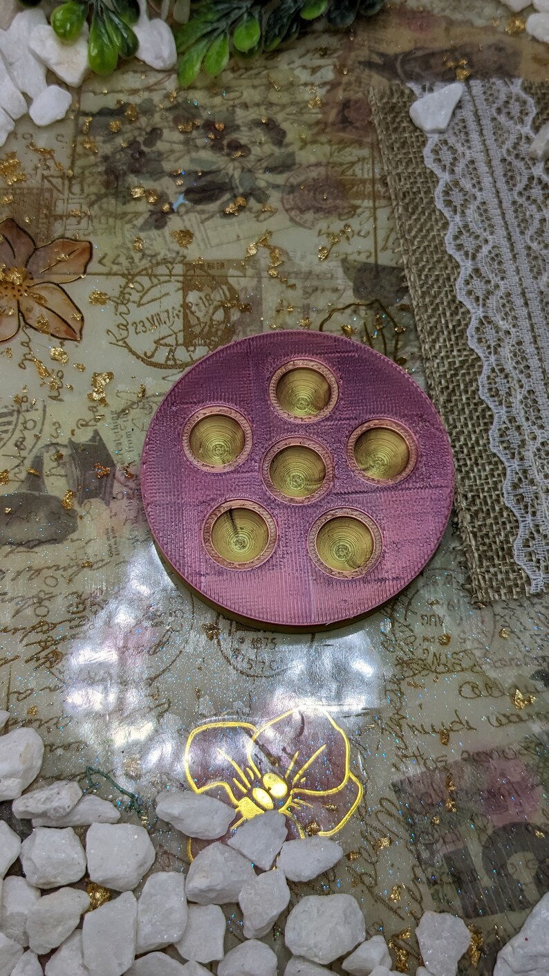 May include: A round, pink, 3D printed tray with six circular indentations. The indentations are gold in colour and have a concentric circle design.