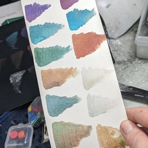Pu&ograve; includere: Un foglio di carta bianco con 14 campioni di acquerello in vari colori, tra cui verde, viola, blu, rosso, marrone e grigio. La carta &egrave; su una bilancia nera con la parola "TARE" stampata su di essa.