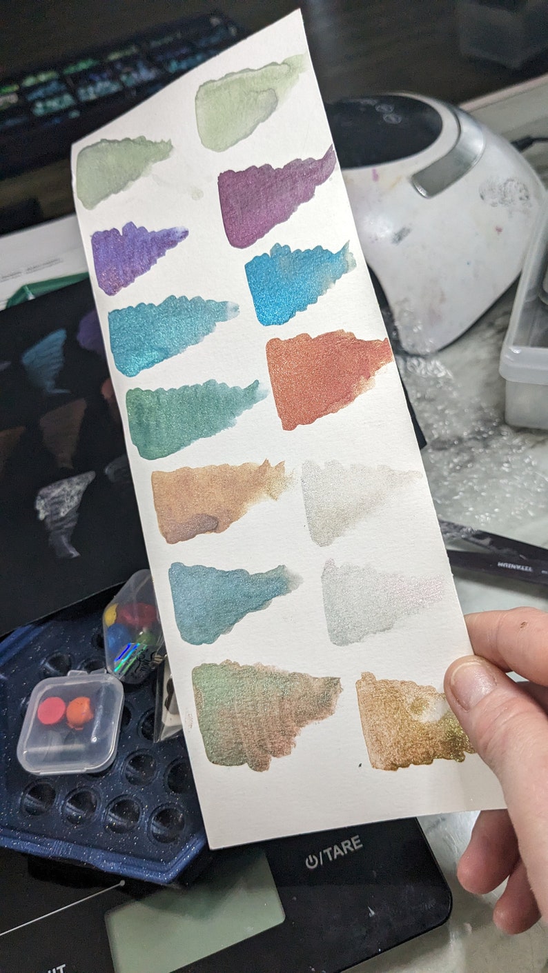 Pu&ograve; includere: Un foglio di carta bianco con 14 campioni di acquerello in vari colori, tra cui verde, viola, blu, rosso, marrone e grigio. La carta &egrave; su una bilancia nera con la parola "TARE" stampata su di essa.