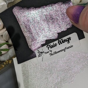 May include: A sample of iridescent glitter paint called "Pixie Wings" by The Blooming Faerie. The paint is a light purple with a silver shimmer and is shown on a black background and a white background.