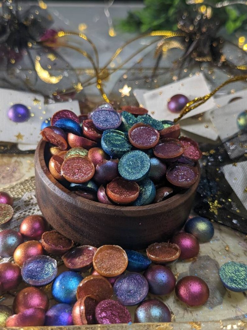 Pu&ograve; includere: Una ciotola di legno piena di cabochon rotondi, piatti e iridescenti di vari colori. La ciotola &egrave; circondata da altri cabochon in varie tonalit&agrave; di blu, verde, viola e arancione.