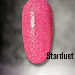 May include: A pink nail polish swatch with glitter, labeled "Stardust" against a dark background. The words "Bloom Dip" are written in pink script at the top. The bottom of the image shows a water reflection.