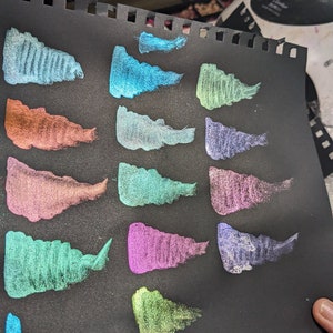 Pu&ograve; includere: Un foglio di carta nero con 16 campioni di vernice metallizzata in vari colori, tra cui verde, blu, viola e arancione. I campioni sono disposti in due file di otto.
