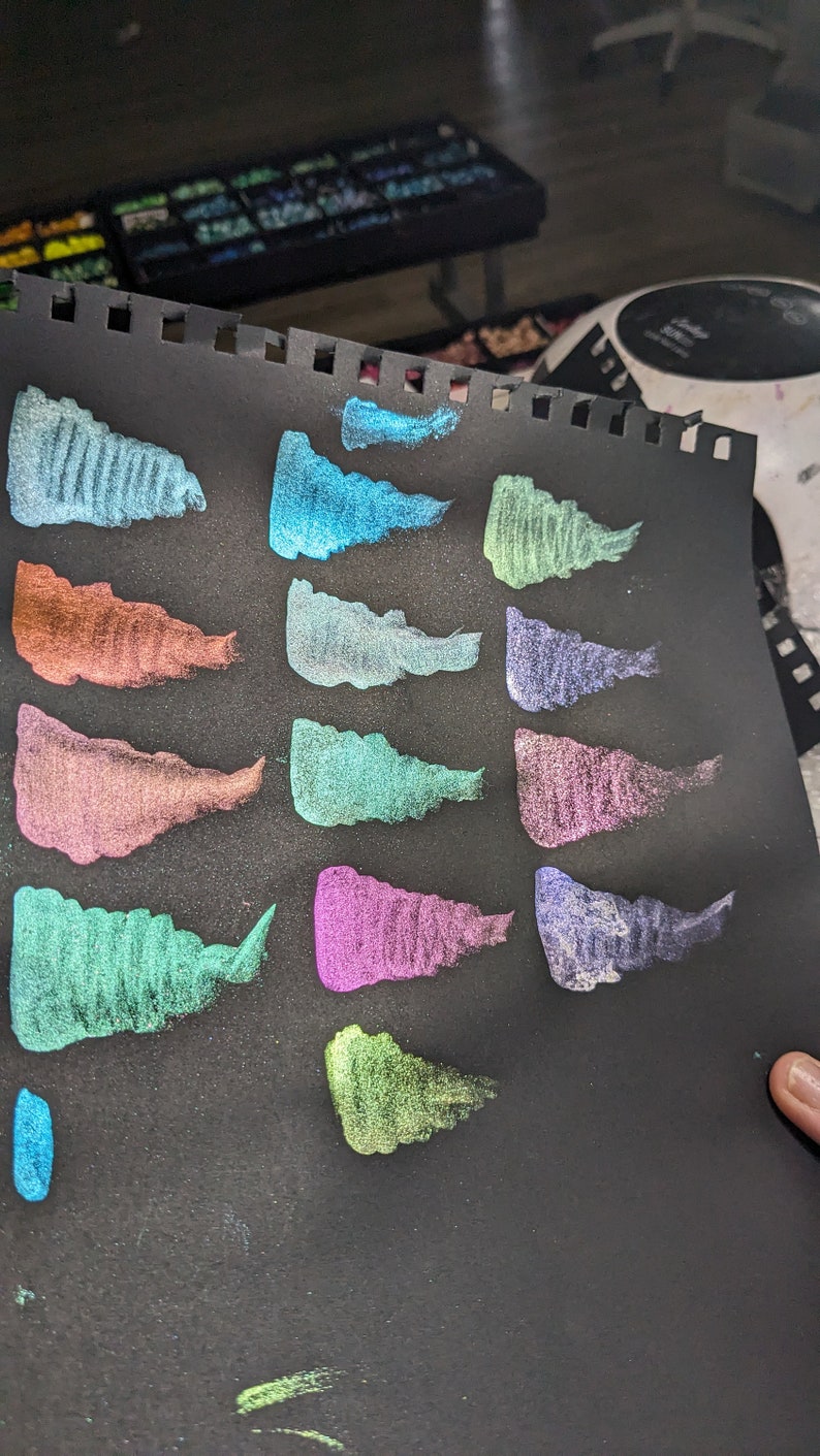 Pu&ograve; includere: Un foglio di carta nero con 16 campioni di vernice metallizzata in vari colori, tra cui verde, blu, viola e arancione. I campioni sono disposti in due file di otto.