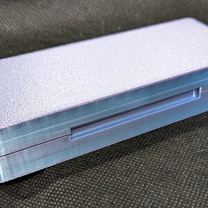 May include: A small, rectangular, light purple box with a textured surface. The box has a thin, light blue, rectangular slot on the side.