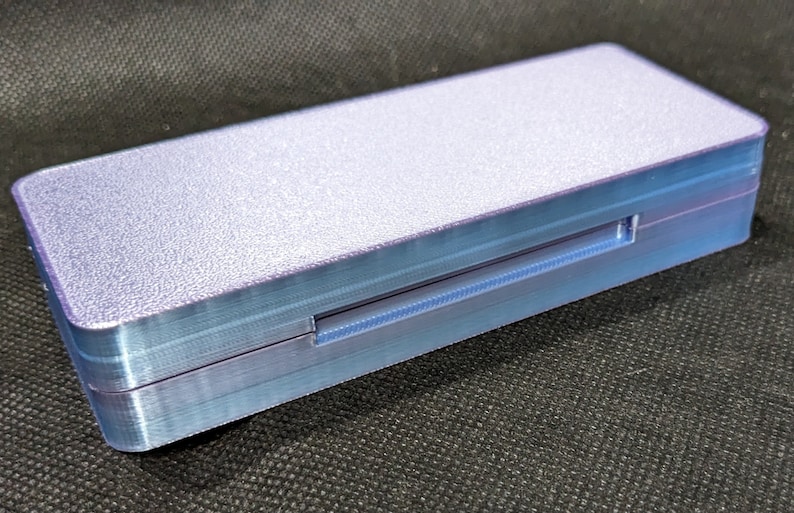 May include: A small, rectangular, light purple box with a textured surface. The box has a thin, light blue, rectangular slot on the side.