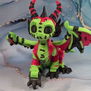 May include: A 3D-printed dragon figurine in a vibrant color scheme. The dragon features a green body with red accents on the chest and wings. Black details include horns, claws, and wing supports. The dragon has a stitched-mouth design.
