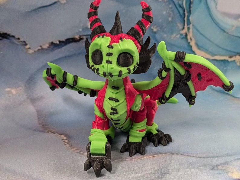 May include: A 3D-printed dragon figurine in a vibrant color scheme. The dragon features a green body with red accents on the chest and wings. Black details include horns, claws, and wing supports. The dragon has a stitched-mouth design.