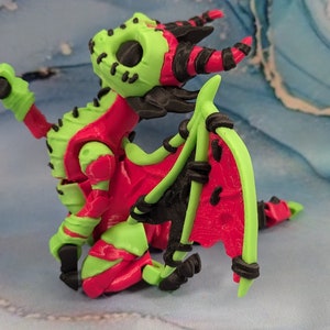 May include: A 3D-printed dragon figurine in vibrant colors. The dragon features a lime green head, limbs, and tail, contrasted by a red body and wings. Black accents highlight the horns, claws, and wing details. The dragon is posed on a blue and white surface.